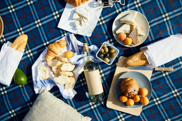 野餐的高角度视图 包括酒瓶 羊角面包 三明治 奶酪和放在格子毯子上的水果 — 图库照片
