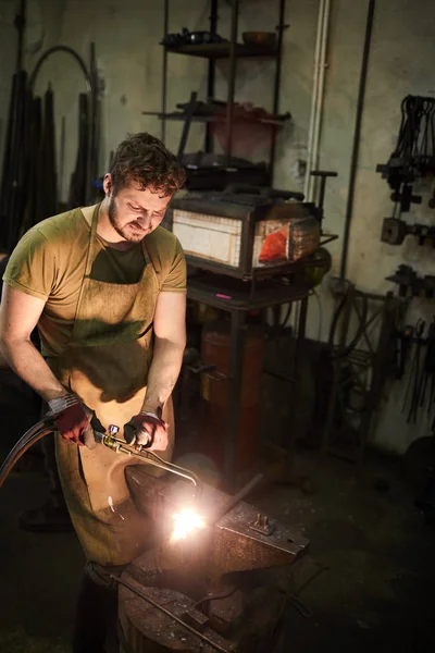 铁匠围裙和手套中的年轻人在石灰中加工铁件时使用焊接工具 — 图库照片