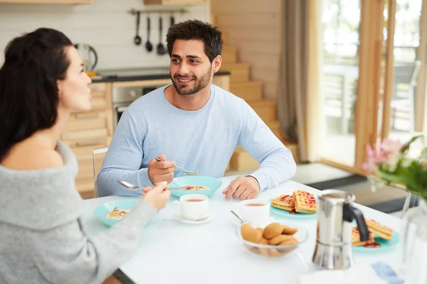 快乐积极的年轻多民族夫妇穿着家庭服装坐在餐桌旁 一边吃健康的早餐 一边在家聊天 — 图库照片