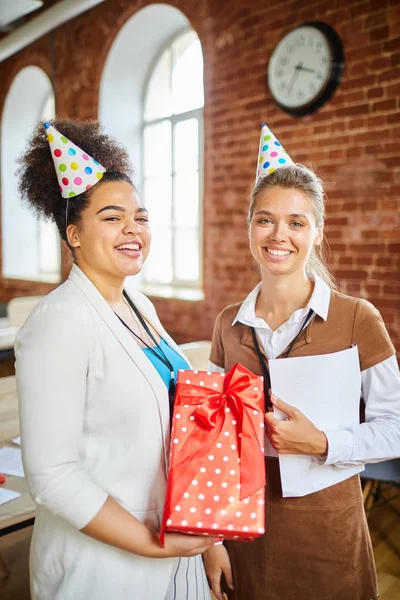 誕生日キャップとカジュアルウェアを着た2人の陽気な若い女性が お祝いの間にオフィスであなたを見ています — ストック写真