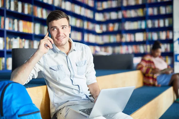 Student Rozmawia Telefon Komórkowy Siedząc Laptopem Bibliotece Kolegium — Zdjęcie stockowe