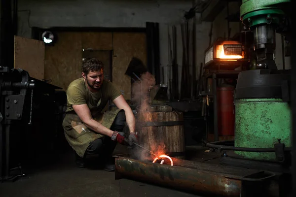 穿着工作服的年轻铁匠把炽热的熔融金属工件放入水中冷却 — 图库照片