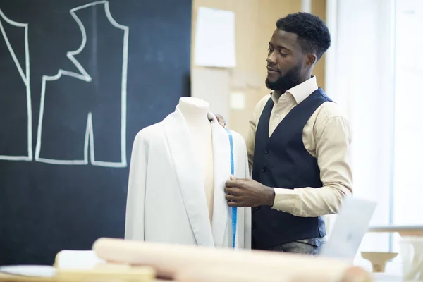 认真沉思英俊的年轻非洲裔美国裁缝与胡子站在假人 并采取夹克措施 而工作夹克设计 — 图库照片