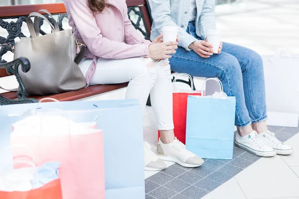 两个穿着牛仔裤和夹克的女孩坐在长凳上 购物后喝酒 讨论她们的购买情况 — 图库照片