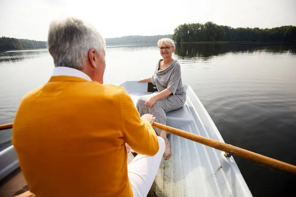 Happy attractive elderly lady in dress and glasses sitting on boat and looking at man while they riding boat during beautiful date on lake