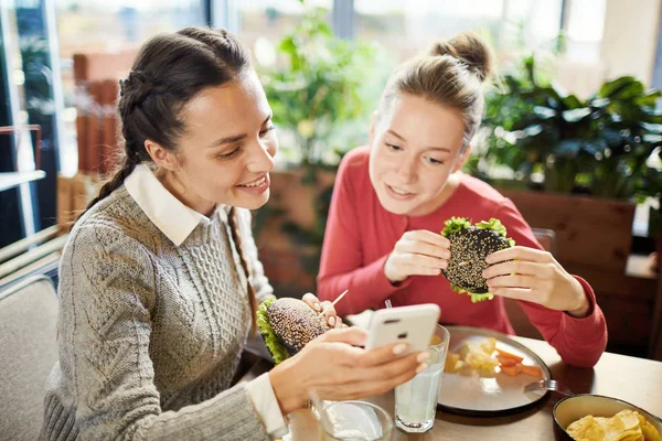 チーズバーガーを食べ Cfaeで昼食でスマートフォンでビデオを見ている2人のフレンドリーな女の子 — ストック写真