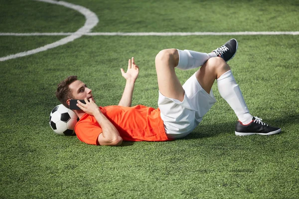 サッカー ボールの頭と緑の野原に横たわって 誰かと話はスマート フォンで若いフットボール選手 — ストック写真