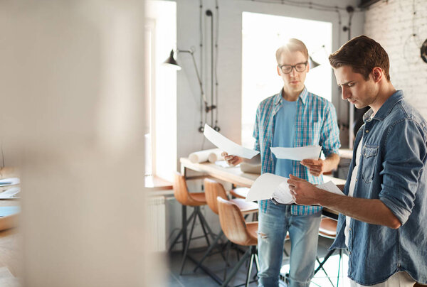 Портрет двух молодых людей в повседневной одежде, работающих в современном офисе, акцент на красивом мужчине, читающем документ на переднем плане, копирующем пространство
