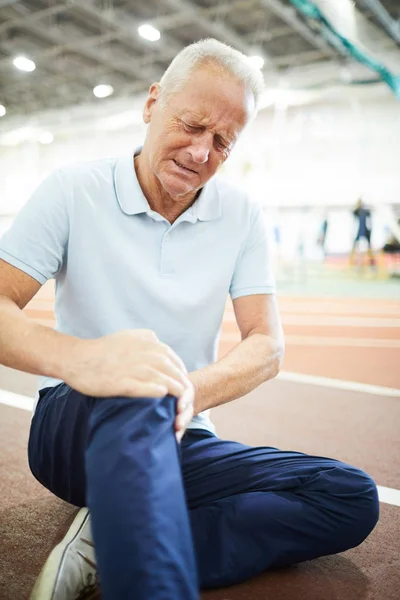 Active senior man sitting on sports track at stadium with his hand on broken knee