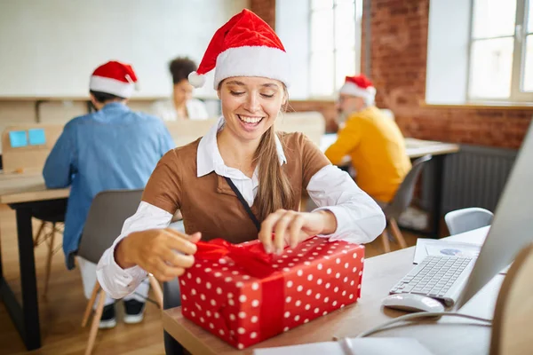 同僚へのプレゼントを準備しながら ギフトボックスの上に赤いシルクのリボンを結ぶ幸せなクリスマスの女の子 — ストック写真