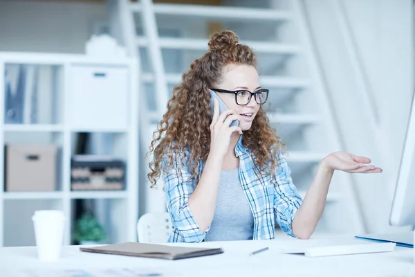 Młoda Bizneswoman Falowanymi Włosami Rozmawia Przez Smartfona Siedząc Przy Monitorze — Zdjęcie stockowe