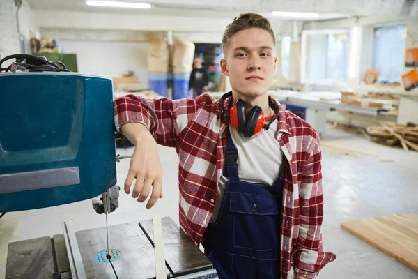 Serious handsome young carpenter with ear protectors on neck leaning on drilling machine and looking at camera in workshop