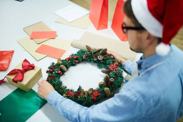 ギフトボックスとカラフルな紙でテーブルに立ち 現代のワークショップでクリスマスリースを飾る忙しい若者 — ストック写真