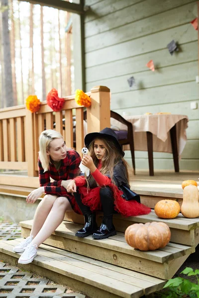 Young blonde woman in flannel dress and her little daughter in witch attire sitting on staircase by terrace of their country house