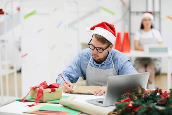 Серьезный Концентрированный Молодой Дизайнер Санта Шляпе Сидит Столом Ноутбуком Подарочной — стоковое фото