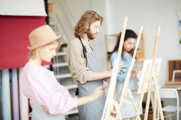 Young man in apron standing by easel between groupmates and painting at lesson in art studio