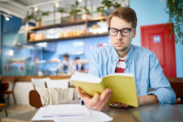 Серьезный Молодой Экономист Сидит Столом Читает Книгу Финансовыми Бумагами Рядом — стоковое фото