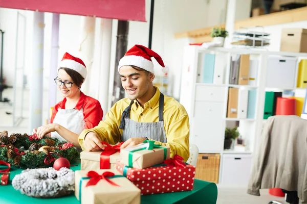 エプロンの若い男とサンタキャップパッキングクリスマスプレゼントと近くの同僚とそれらを結ぶ — ストック写真