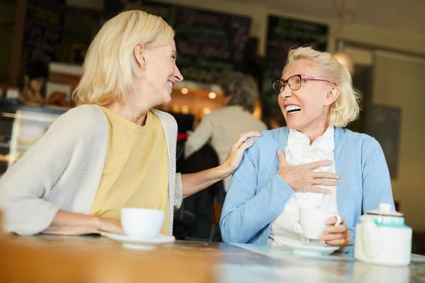 Two female friends laughing during their talk by cup of tea while relaxing in cafe at leisure