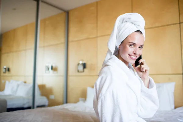 愉快的年轻女子用毛巾在头坐在卧室或酒店房间 通过智能手机说话 看着你 — 图库照片