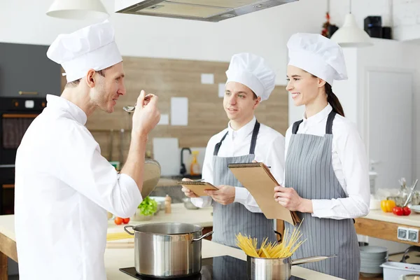 若い料理人と経験を分かち合いながら 料理教室で水で塩を味わう帽子と制服姿のシェフを見る陽気なポジティブな学生たち — ストック写真