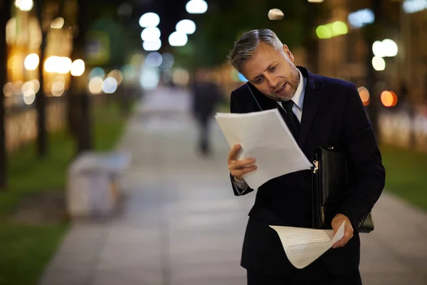 Very busy banker talking by smartphone and looking through financial documents in urban environment after work