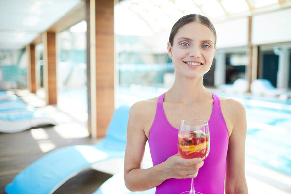 Young fit woman with glass of cocktail looking at camera with row of deckchairs on background