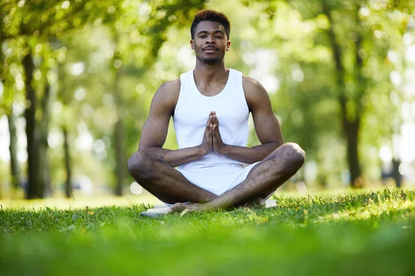 Content handsome young black man with mustache sitting with crossed legs and holding hands in Namaste while practicing yoga in summer park