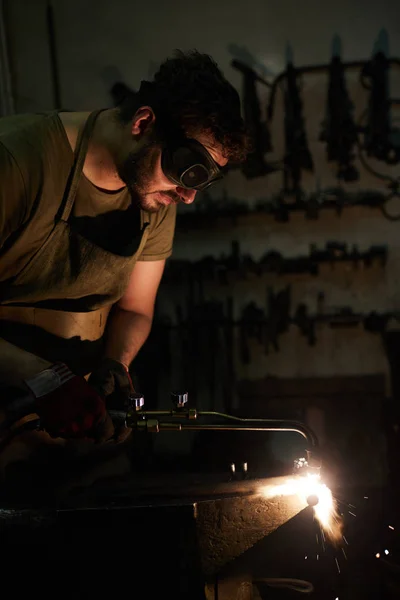 严重集中的胡须男子在深色护目镜使用切割割炬 同时在黑暗的车间切割钢细节在铁锤 火花吹出 — 图库照片