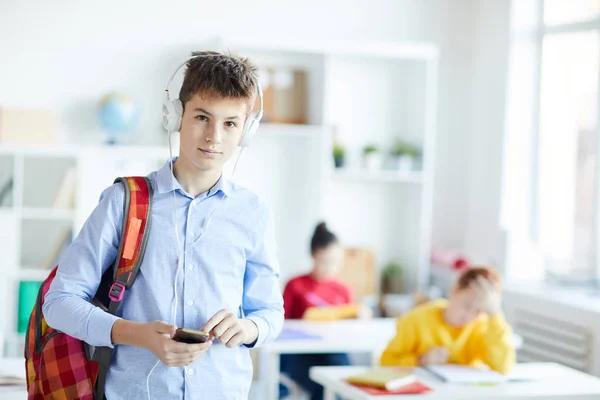 カメラの前に立つバックパック スマートフォン ヘッドフォンを持つ現代の男子生徒 — ストック写真