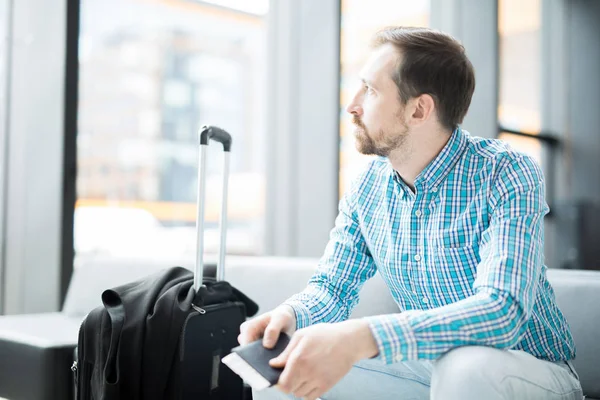 若いビジネスマンは出発のお知らせを待っている間空港ラウンジで彼の手荷物に座って旅行中 — ストック写真