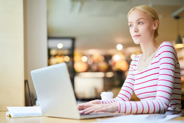 穿着条纹套头衫的年轻 沉思的金发女子坐在笔记本电脑前的咖啡店里 执行任务 — 图库照片