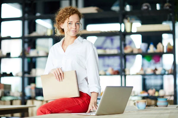 Young creative student of art course sitting on table by laptop in modern studio of crafts