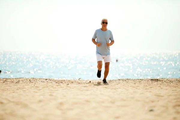 Active pensioner in sportswear running down sandy beach with water on background