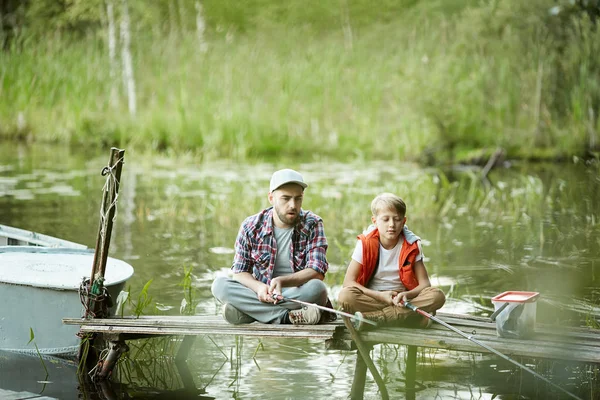 父亲和儿子坐在鱼架上 他们的棍子在水中等待鱼 — 图库照片