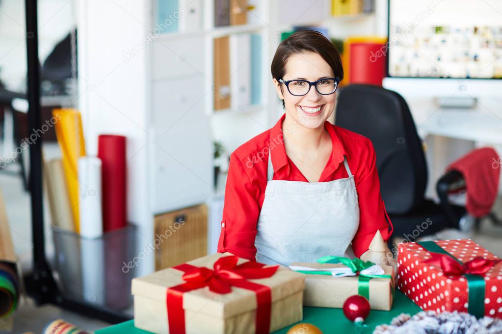 Cheerful young female in red blouse and apron looking at you while sitting by table with gifts