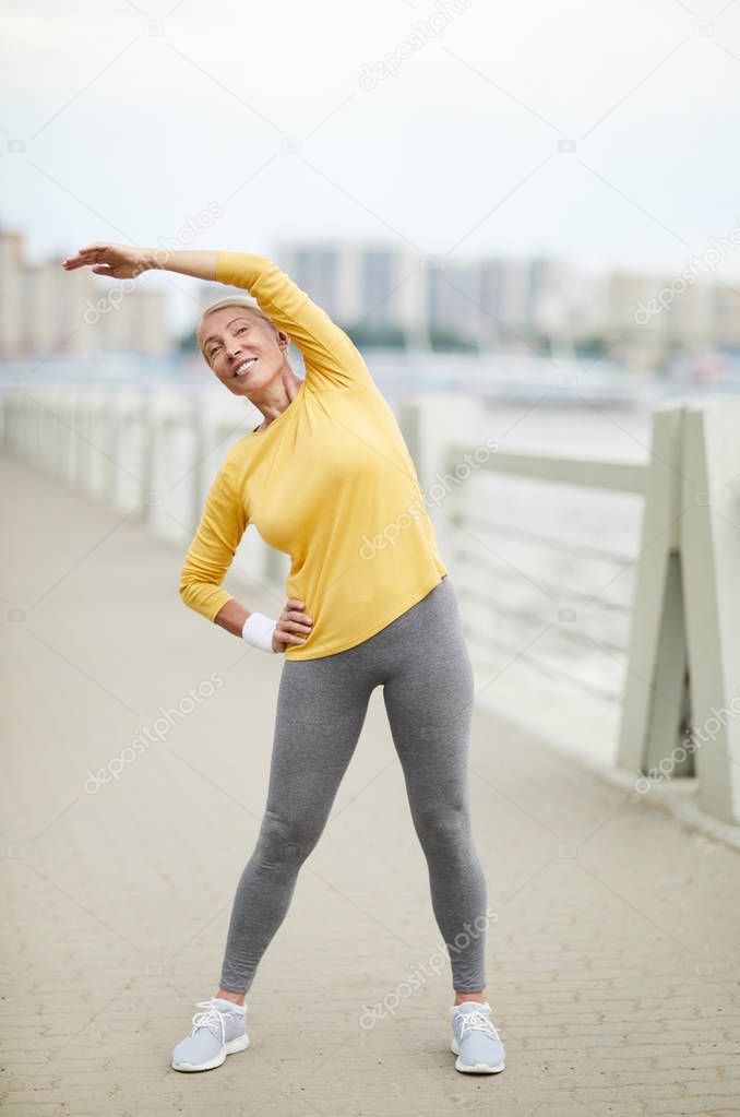 Active mature female in pullover and leggins doing side-bends in urban environment by riverside