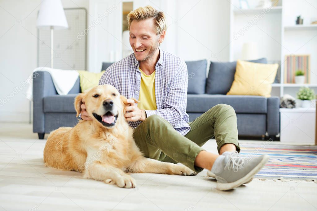 Cheerful young man in casualwear relaxing at home and playing with friendly purebred golden labrador
