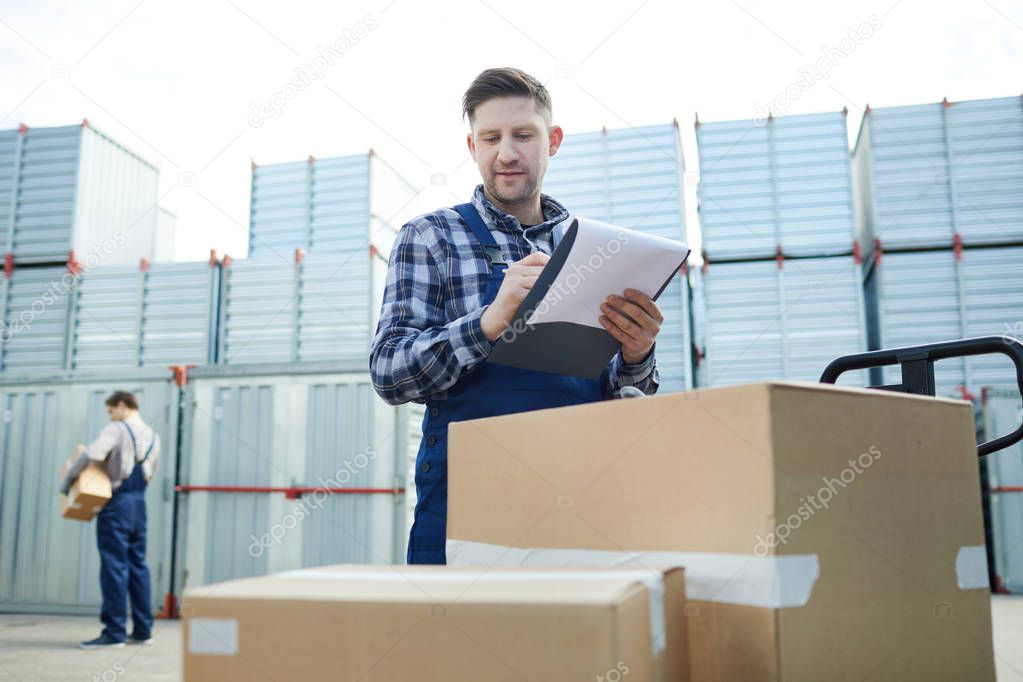 Content confident handsome male cargo distribution worker checking boxes and making notes in document while standing at pullet jack in container storage area