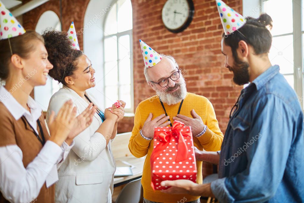 Two happy businesswomen clapping their hands while young man giving birthday present to senior boss
