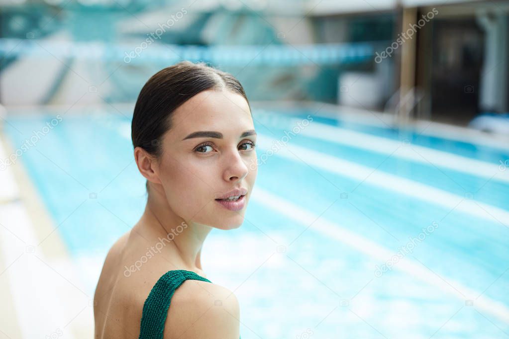Young female looking at you while standing on background of swimming-pool with blue water
