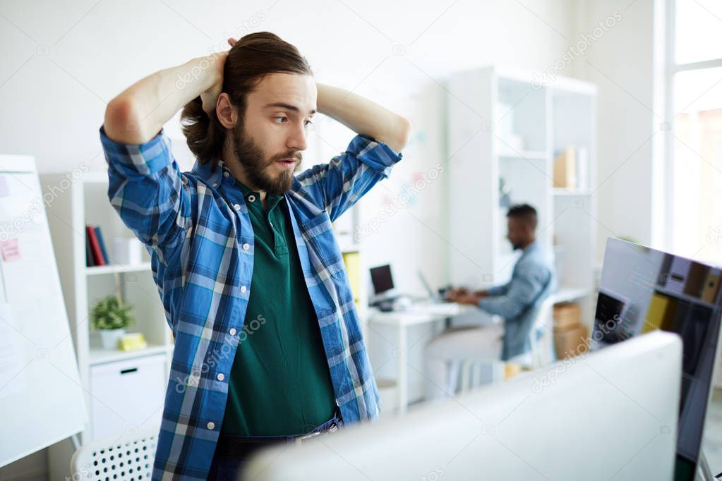Anxious young it-manager touching his head while looking at coded data on one of monitors