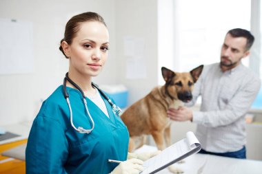 Young serious veterinarian making notes in document and looking at you on background of dog and its owner
