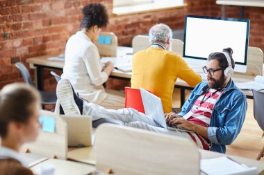 Genç yönetici ya da günlük giyim ve kulaklık editörü dizlerinin üzerinde dizlerinin üzerinde oturmuş internette geziniyor ya da online video izliyor.