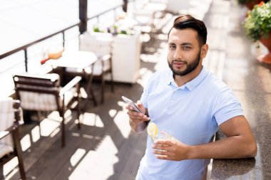 Akıllı telefonu ve bir bardak limonatası olan genç adam açık kafede güneşli bir günün tadını çıkarıyor.