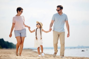 Yaz tatili sırasında sahilde yürüyüş yaparken el ele tutuşan modern mutlu ailenin tam uzunlukta portresi