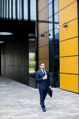 İş elbiseli, gözlüklü, ofis bloğuna yakın bir yerde plastik bardak tutan yakışıklı bir erkek.