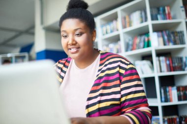 Kütüphanede sınava hazırlanırken neşeli kendine güvenen genç Afrikalı-Amerikalı kilolu öğrenci dizüstü bilgisayarda online makale okuma