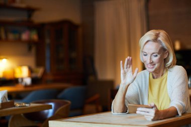Zarif olgun sarışın kadın video-sohbet smartphone içinde aracılığıyla konuşurken elini sallayarak