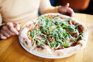 Aç kişinin önünde tabakta taze otlar ve pastırma ile iştah açıcı pizza